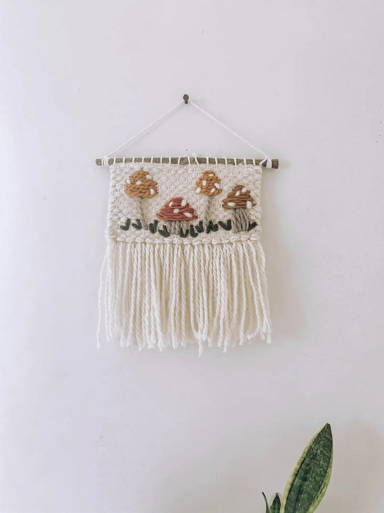 A handmade Mushroom wall hanging featuring mushrooms and foliage designs, with long, dangling fringes, perfect as nursery decor on a white wall.