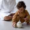 A toddler in a mustard cardigan plays with a Medical Grade Silicone Baby Bottle, sitting on the floor. An adult in white, partially visible, sits nearby, out of focus.