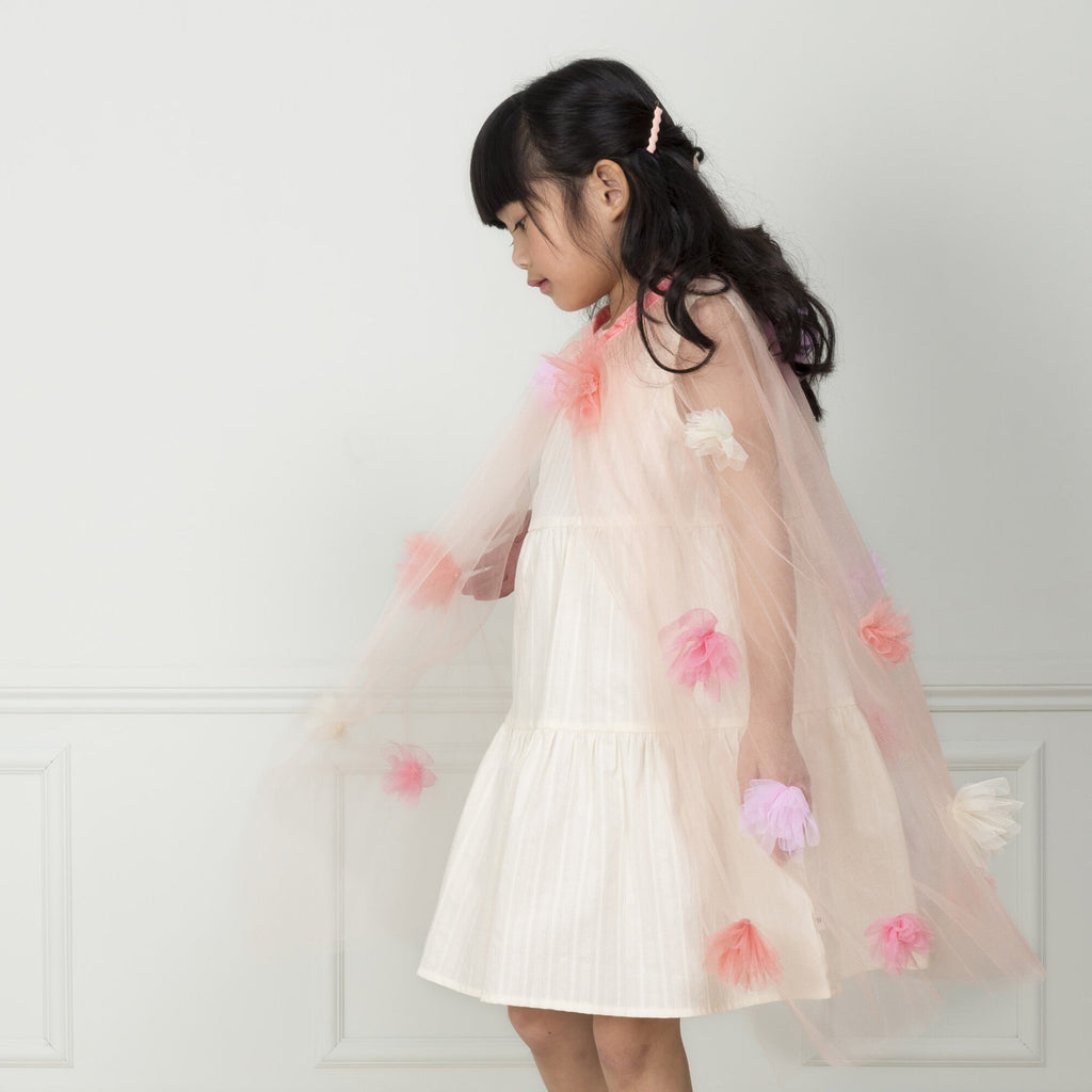 A young girl in a white dress and a Meri Meri Flower Cape twirls in front of a white background, looking down and to her side with a playful expression.