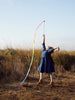 A young girl in a blue dress and white leggings is outside in a grassy field, reaching up to shoot Sarah's Silk Rainbow Streamer Wand from a bow, against a clear sky.