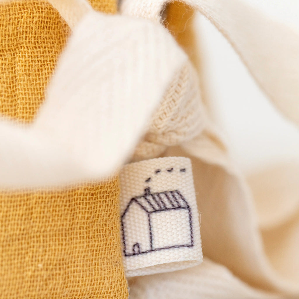 Close-up of a clothing label featuring a small embroidered house design, surrounded by soft, textured organic cotton fabric in shades of white and gold.