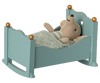 A plush teddy bear lying cozily under a blanket in a Maileg Miniature Cradle with golden knobs.