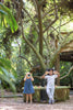Two children playing in a lush garden, one taking a photo with a Wooden Water Blocks camera made sustainably, the other pretending binoculars with doughnuts. Light strikes through the canopy above.