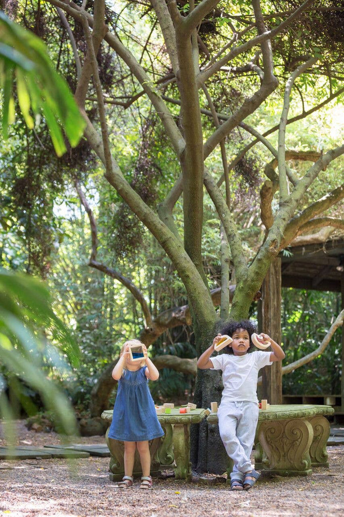 Two children playing in a lush garden, one taking a photo with a Wooden Water Blocks camera made sustainably, the other pretending binoculars with doughnuts. Light strikes through the canopy above.