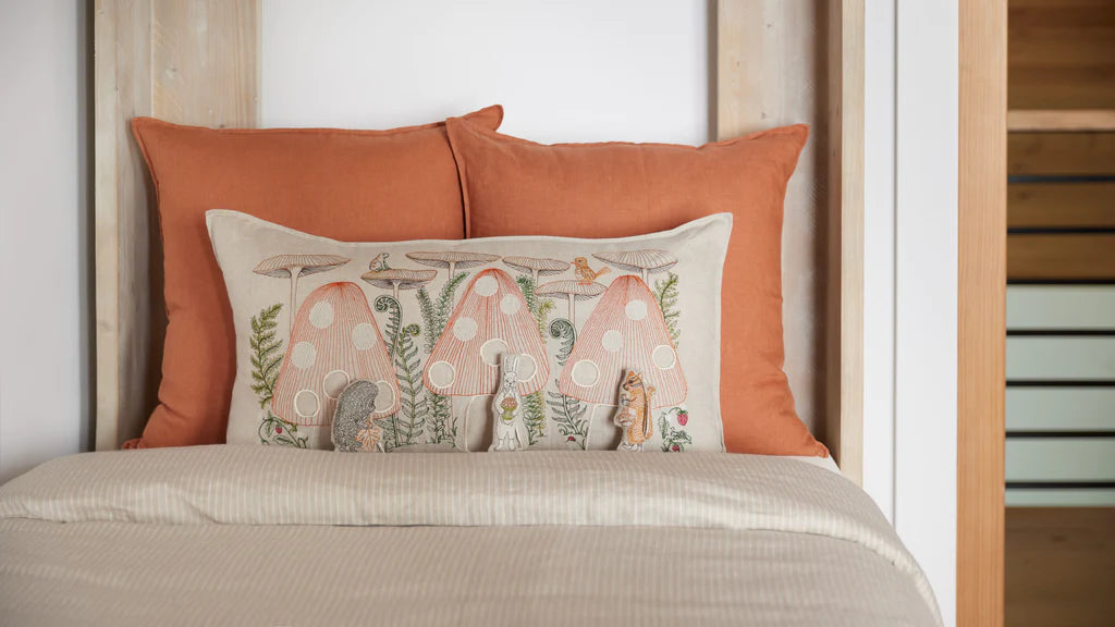 A cozy bedroom scene featuring a neatly made bed with a beige cover and two orange pillows with one Coral & Tusk Mushroom Forest Pocket Pillow in front.