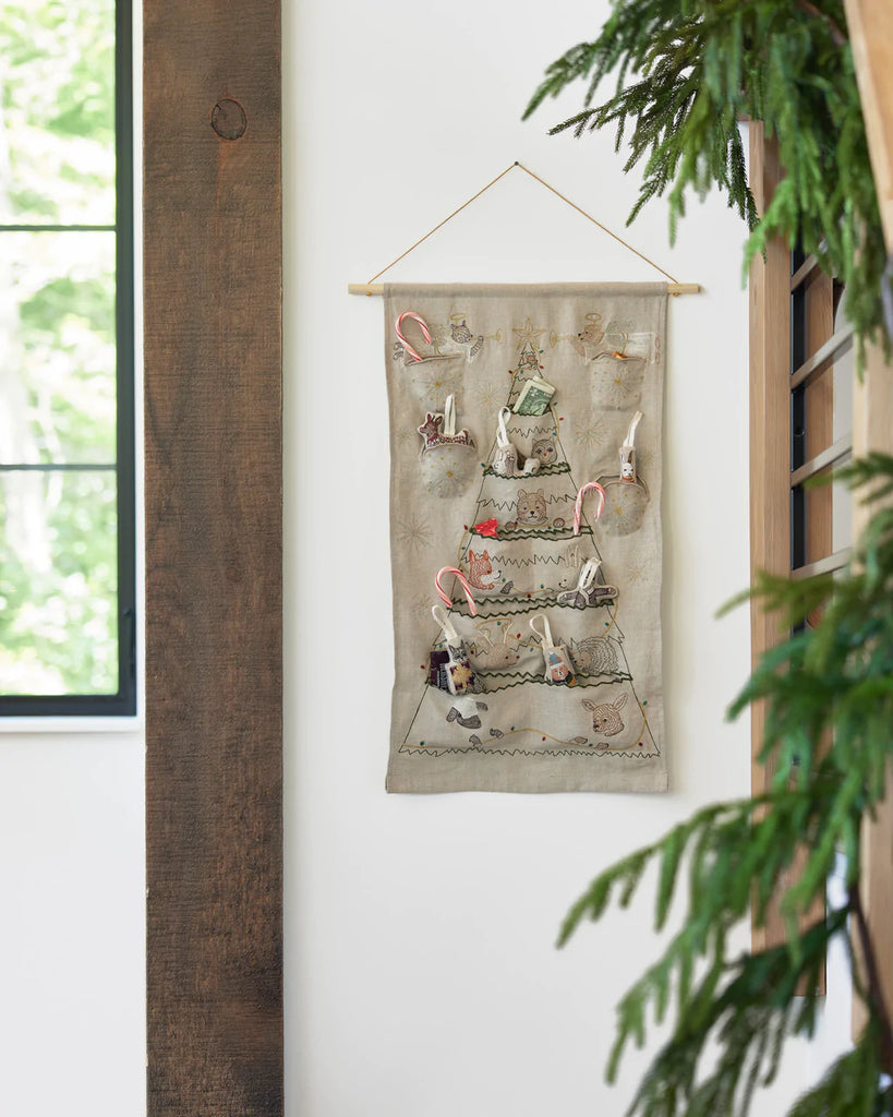 A Coral & Tusk Embroidered Advent Calendar hangs on a white wall between a window and a wooden beam, decorated with festive ornaments and pockets for small gifts.
