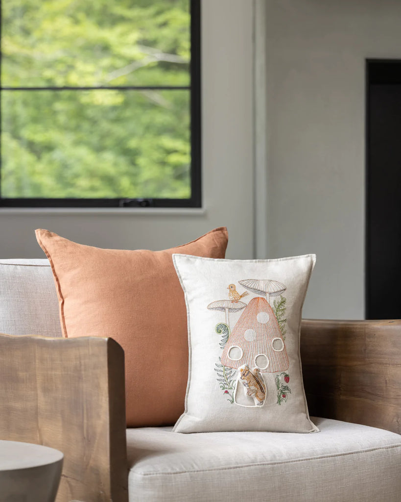 A Coral & Tusk Mushroom House Pocket Pillow with an embroidered mushroom house design sits on a beige armchair, flanked by a plain brown pillow, with a view of green trees through a nearby window.
