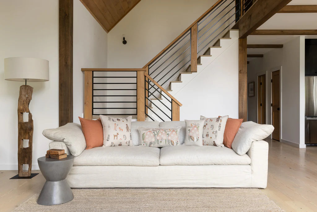 A cozy, modern living room featuring a Coral & Tusk Mushroom House Pocket Pillow with decorative pillows, a wooden floor lamp, and a staircase with metal and wood railings in the background.