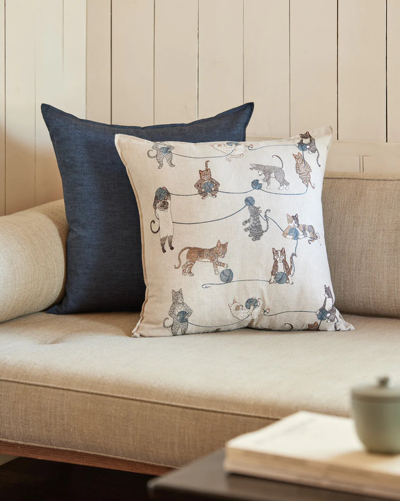 Decorative embroidered Coral & Tusk Playful Cats pillow with Siamese cat illustrations on a beige sofa next to a dark blue pillow, placed against a white paneled wall. A light tea cup is also visible on the sofa.