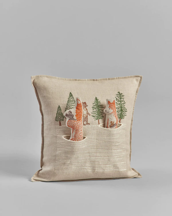 A Coral & Tusk Winter Foxes Pocket Pillow featuring embroidered woodland animals, including a fox hunt in snow, and trees on a gray background.