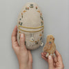 A person holding a Coral & Tusk Pocket Easter Egg decorated with animal motifs, alongside a small embroidered bird, set against a neutral background.