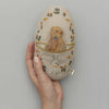 A hand holding a Coral & Tusk Pocket Easter Egg with an embroidered design featuring a bird on a swing and a bunny surrounded by small flowers, set against a light gray background.