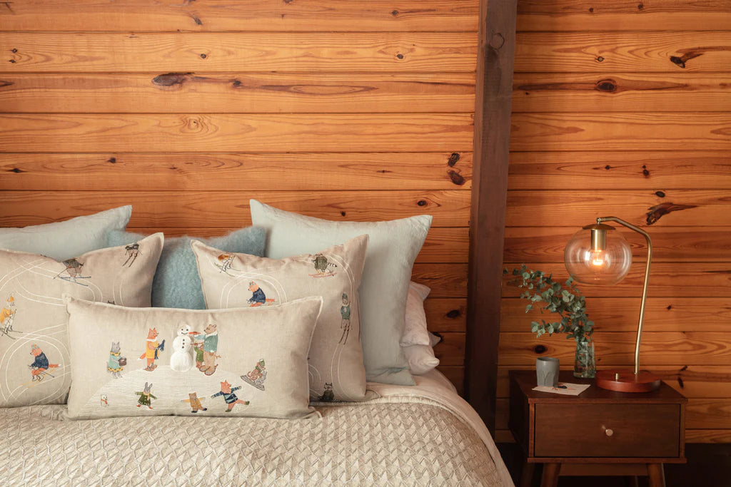 A cozy bedroom corner featuring a large wooden headboard and bed covered with various pillows, including the Coral & Tusk Downhill Skiers Pillow adorned with embroidery motifs. A wooden nightstand with a lamp and plant sits to the side.