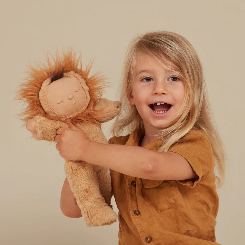 A joyful young child with long blonde hair, wearing a mustard shirt, holds up a Olli Ella Cozy Dinkum Doll - Lion Pip with an embroidered face and a big smile, against a beige background.