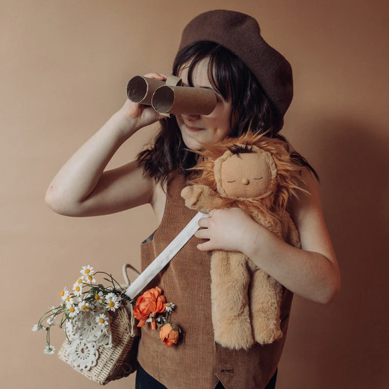 A child wearing a hat looks through a makeshift telescope, holding a Olli Ella Cozy Dinkum Doll - Lion Pip and a flower-filled basket, against a brown backdrop.