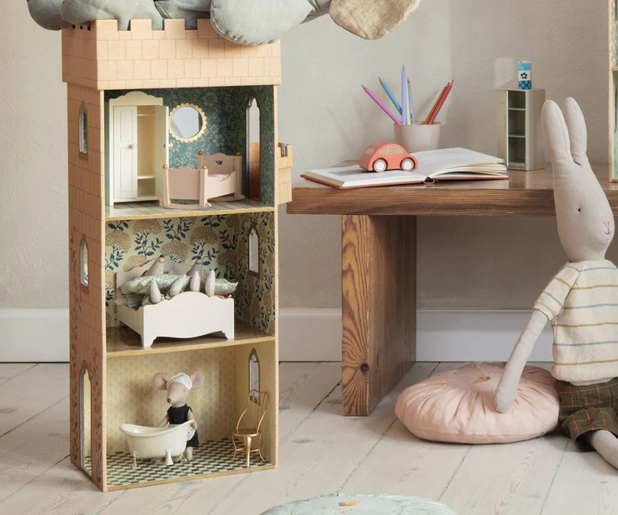 A creatively designed Maileg Miniature Cradle beside a wooden desk, with a stuffed rabbit toy and a mouse set sitting on the floor, a car toy, books, and pencils on the desk.