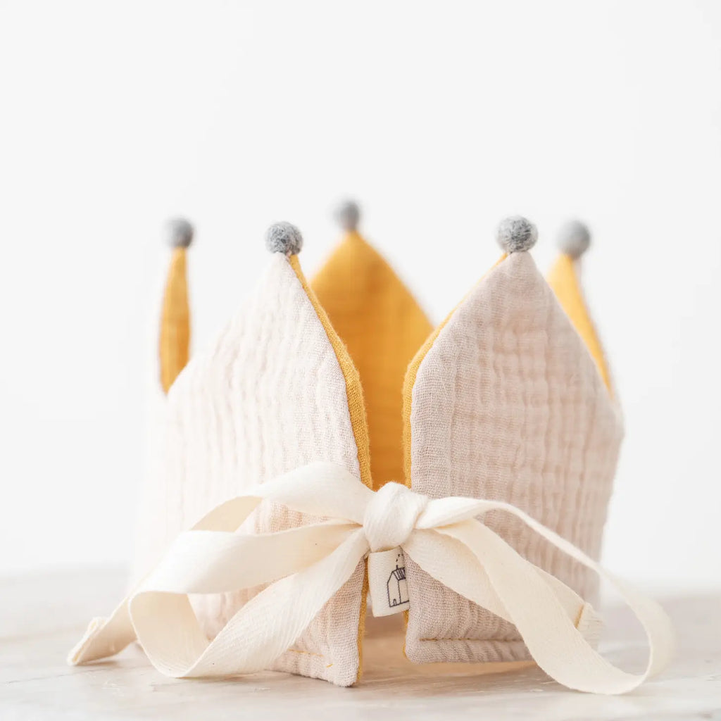 A handmade Reversible Crown crafted from organic cotton with alternating yellow and white segments, adorned with gray pom-poms on top and tied with a cream ribbon, placed on a light surface.