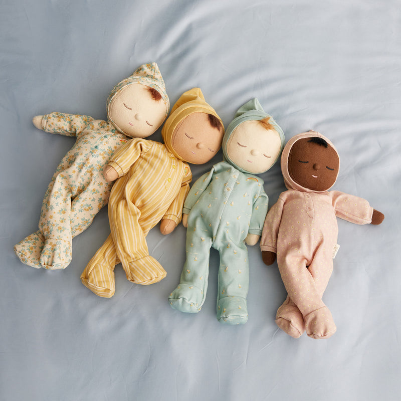 Four diverse Olli Ella Dozy Dinkum Doll - Daydream Edition dolls lying side by side on a light blue fabric surface, each dressed in different patterned 100% cotton pajamas.