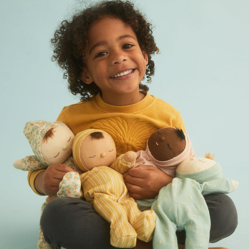 A joyful young child with curly hair, wearing a yellow sweater, holding three limited edition Olli Ella Dozy Dinkum Doll - Daydream Edition dolls and smiling at the camera against a light blue background.