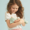A young girl with curly hair gently holding a Olli Ella Dozy Dinkum Doll - Daydream Edition stuffed toy, smiling down at it affectionately. She is wearing a white t-shirt and pink skirt against a soft blue landscape.