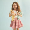 A smiling young girl with curly hair holding two stuffed toys, dressed in a pink ruffled skirt and white top from the limited edition Olli Ella Dozy Dinkum Doll - Daydream Edition collection, standing against a light blue