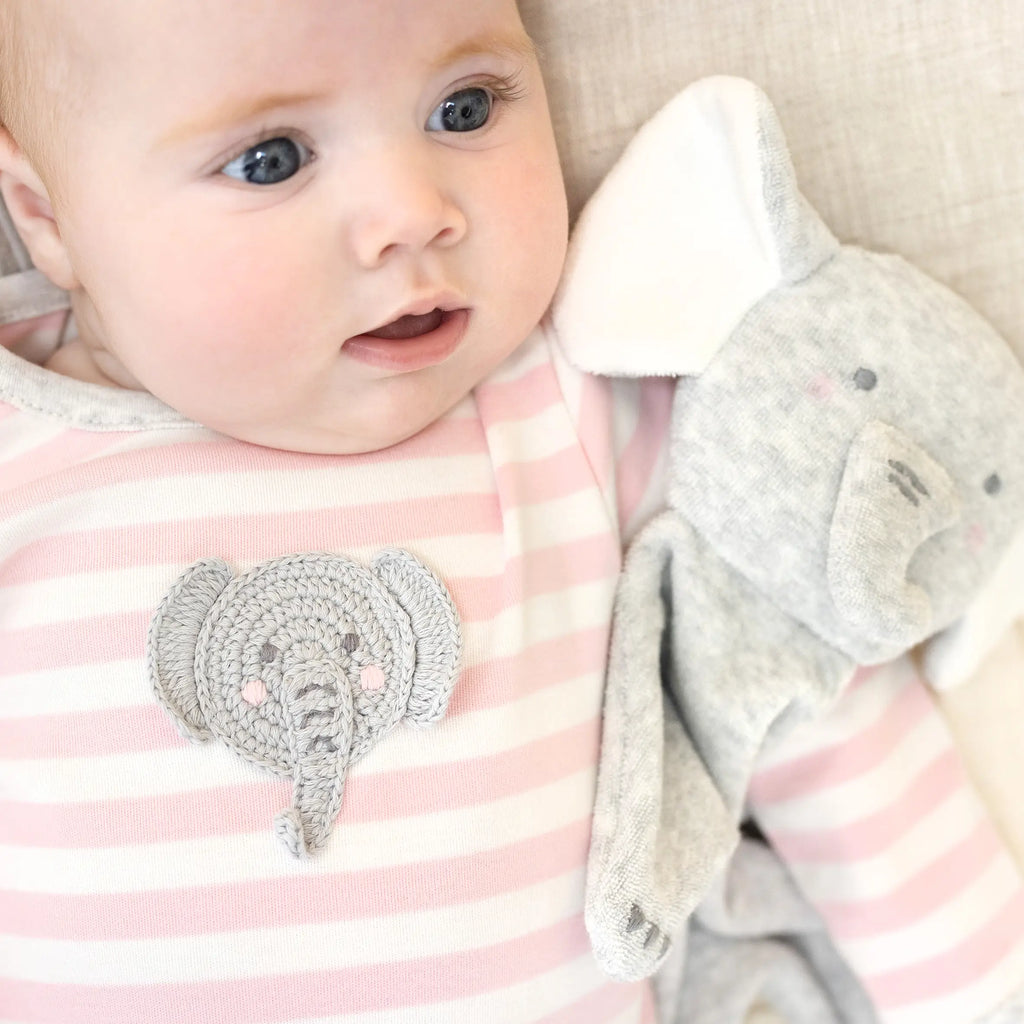 A young baby lying on a soft surface looks curious, wearing a pink and white striped outfit made of organic cotton with an Elephant Lovey design, next to a plush elephant toy.