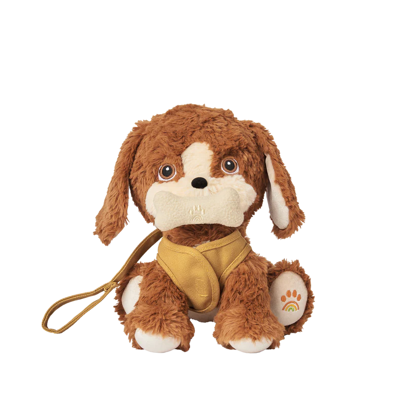A plush toy dog from Olli Ella Dinkum Dogs with brown and white fur, big puppy eyes, and a golden bandana sits against a plain background, holding a magnetic bone accessory in its mouth.