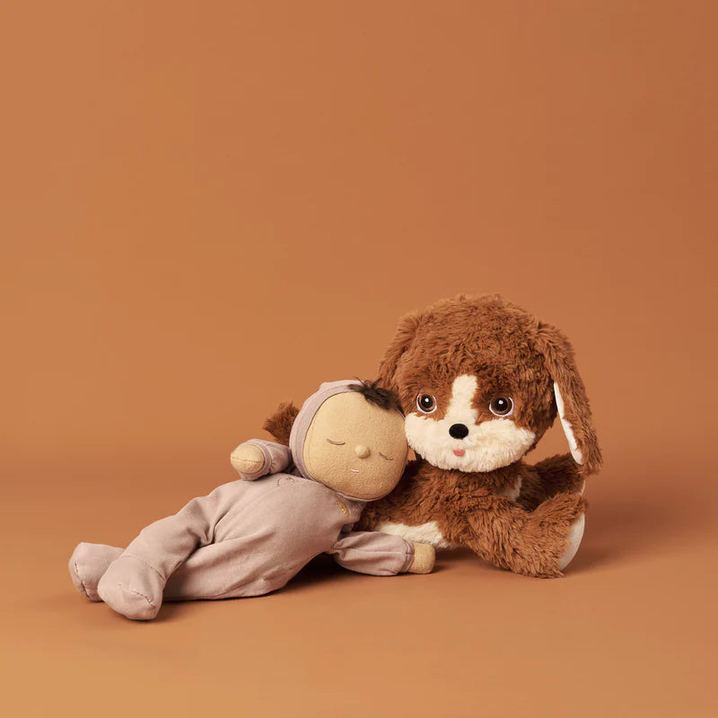 A Olli Ella Dinkum Dog with brown and white plush fabric, sitting next to a rag doll with a beige body and brown hair, both on a plain brown background.