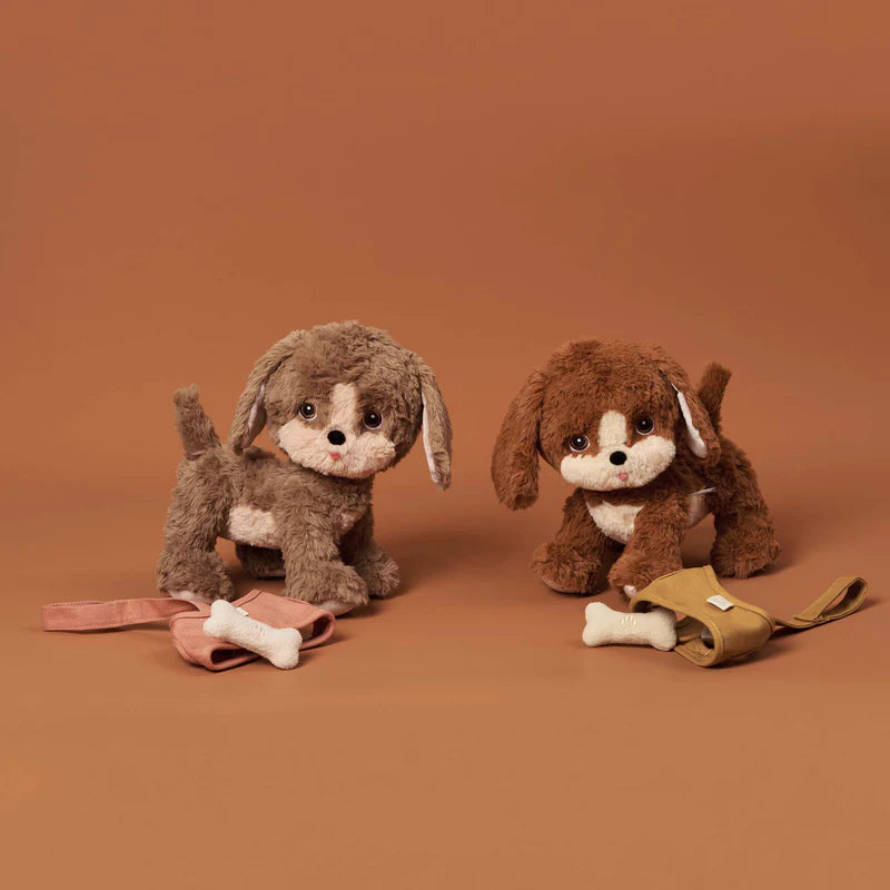 Two Olli Ella Dinkum Dogs, one light brown and one dark brown, sitting side by side against a plain tan background. Each toy has expressive eyes and fluffy fur crafted from plush fabric.