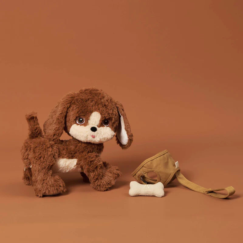 A Olli Ella Dinkum Dog with brown and white fur sits against a matching tan background, with one ear flopped down and a small magnetic bone accessory nearby.