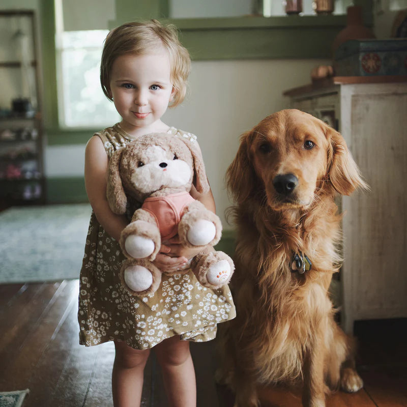 A young girl holding a Olli Ella Dinkum Dog stands next to a golden retriever wearing a magnetic bone accessory inside a cozy room, both looking directly at the camera.