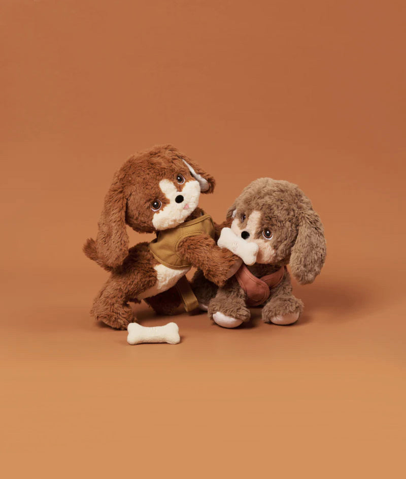 Two Olli Ella Dinkum Dogs, one brown and one beige, are seated against a peach background, closely hugging each other with a magnetic bone accessory nearby.