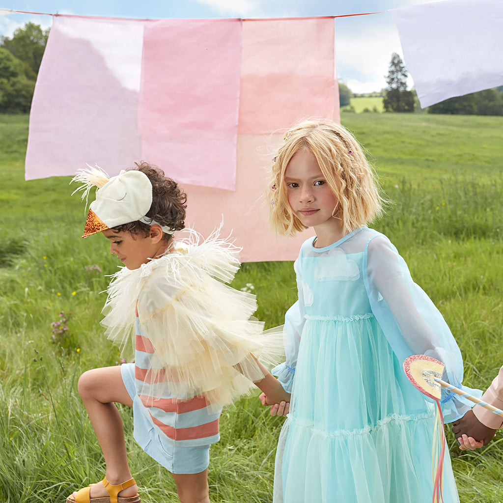 Two children play outdoors near colorful hanging fabrics. One child in a Meri Meri Chick Costume and another in a blue dress, both looking at the camera in a grassy field.