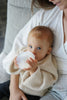 A baby with blue eyes, wearing a beige sweater, is held by a woman and drinking from a white Medical Grade Silicone Baby Bottle. Only part of the woman's face and her arms are visible.