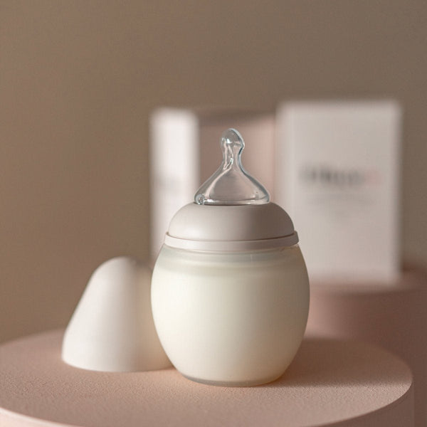 A modern Medical Grade Silicone baby bottle and its lid on a round pink table, with a soft beige background and a boxed product visible behind it. The bottle features a silicone nipple and a minimalist design.