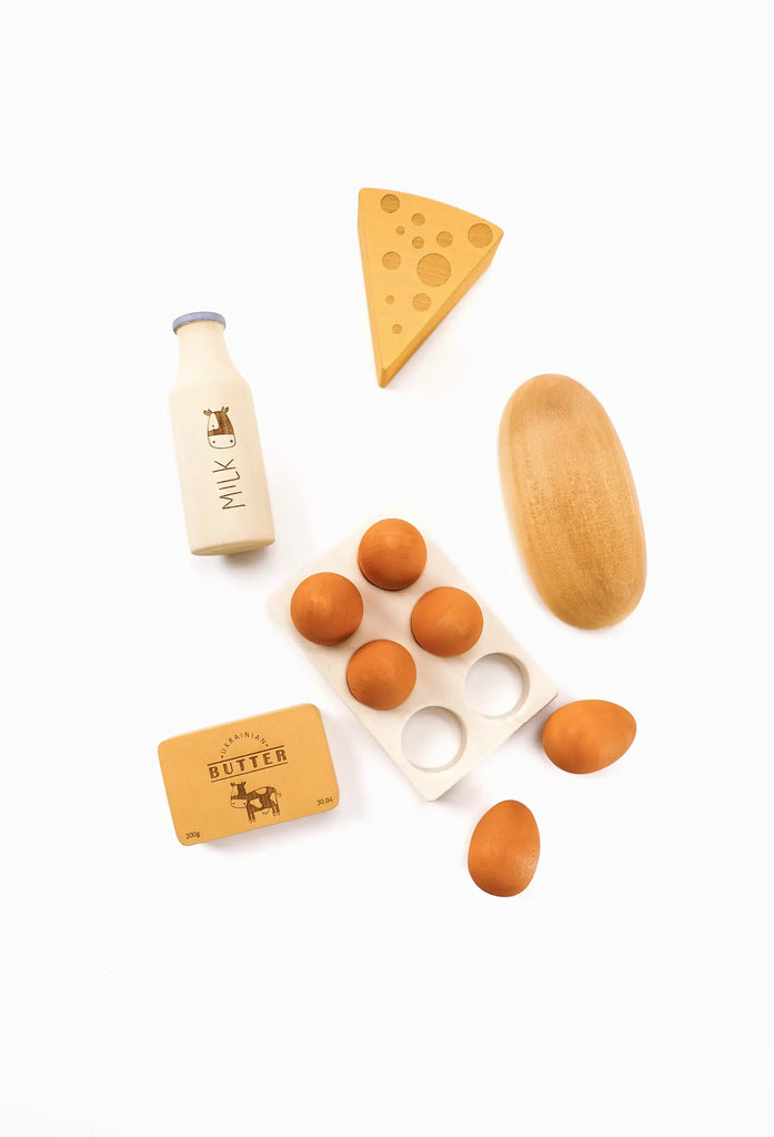 Flat lay photo showing Sabo Concept Handmade Wooden Dairy Set items: a bottle of milk, a triangle of cheese, eggs on a tray, a loaf of bread, and a block of butter, all against a white background.