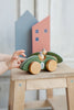 A child's hand reaches for a green Handmade Wooden Toy Race Car with large yellow wheels and two small wooden figures inside. The handcrafted car sits on a wooden stool. In the background, there are two shapes resembling houses, one painted blue and the other red, all made from sustainably harvested birch wood.