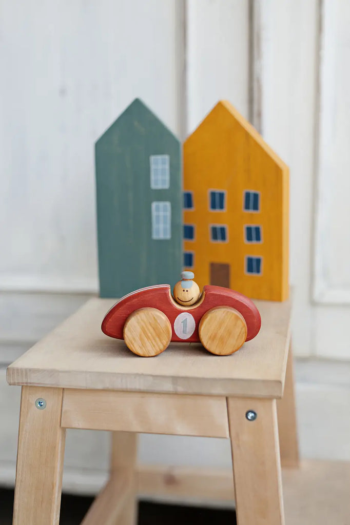 A Handmade Wooden Toy Race Car with a small figure sits on a light-colored wooden stool. Behind it, there are two decorative wooden houses, one painted green with white windows and the other yellow with blue windows, all made from sustainably harvested birch wood.