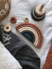 Top view of various wooden toys on a textured white blanket, including a Handmade Rainbow Stacker - Terracotta and rings next to a striped gray cloth and a woven basket. The stacking toy features non-toxic paint, ensuring