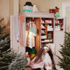 Two children dressed in festive costumes, one as an angel, are playing in front of an open wardrobe filled with holiday-themed items and gifts. Among them is a soft knitted Jingles The Reindeer Toy. The scene is set amidst decorated Christmas trees, creating a whimsical, festive atmosphere.