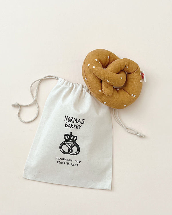 A fabric drawstring bag labeled "norma's bakery" with an eco-friendly screen print, and a handmade plush toy resembling a Handmade Soft Pretzel Play Food next to it on a light background.