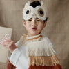 A young child wearing a Meri Meri Owl Costume and a crafted cape stands against a neutral background, puckering lips in a playful expression while holding a piece of paper.