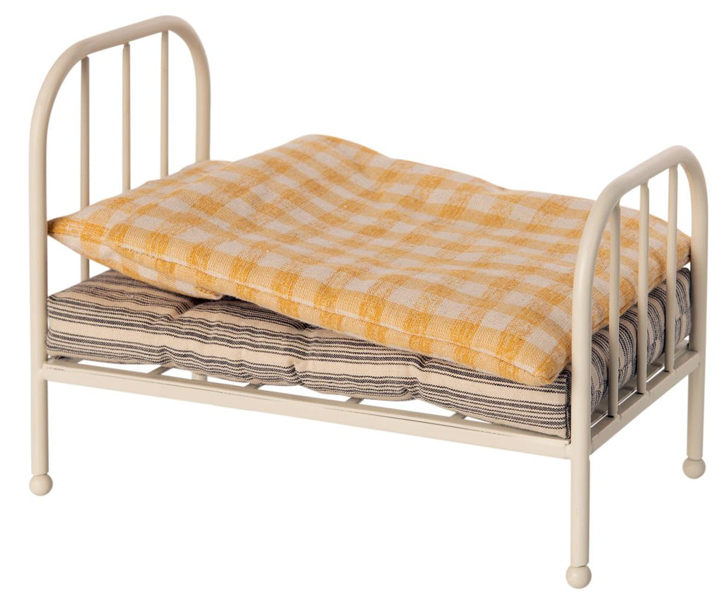 A small Maileg Miniature Bed with a plaid orange and white mattress on top, and neatly folded striped blankets stored underneath, isolated on a white background.