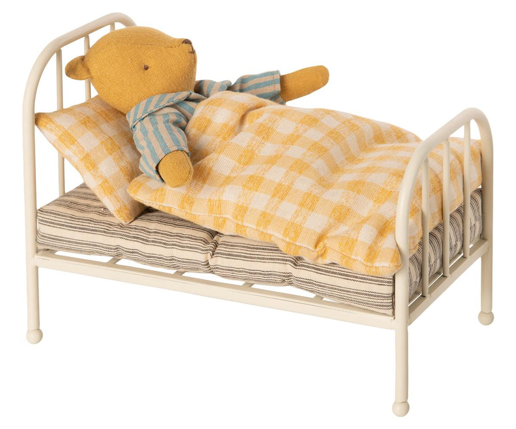 A plush teddy bear lying on a Maileg Miniature Bed covered with a checkered yellow blanket and striped cushions, against a white background.