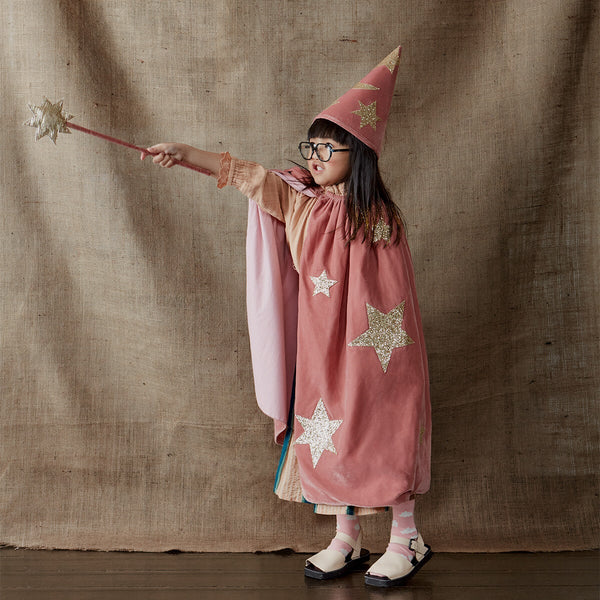 A young child dressed in a Meri Meri Pink Velvet Wizard Costume stands against a textured backdrop, holding a star-tipped wand outstretched. They wear glasses, a pink velvet cape with stars, and a matching conical hat.