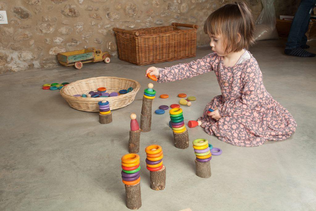 A young girl wearing a floral dress plays with colorful Grapat Nins Carla (Nins, Rings & Coins in 12 Colors) wooden stacking toys on a concrete floor, focusing intently on placing a piece.