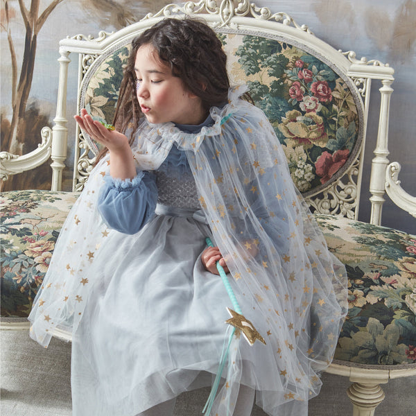 A young girl in a Meri Meri White Tulle Star Cape Costume sits on an ornate chair, blowing a kiss towards her hand, with a painted scenic backdrop.