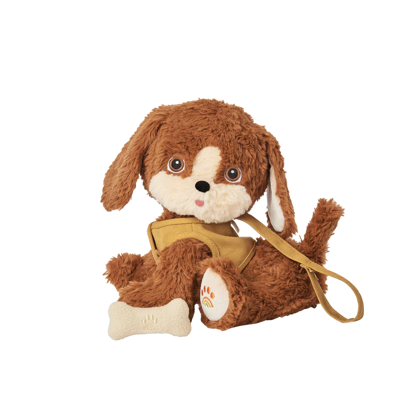 A Olli Ella Dinkum Dog toy with brown fur, large white and black eyes, and a cute expression, wearing a yellow backpack, set against a transparent background.