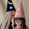 Two children wearing festive cone-shaped party hats, one pink and one navy blue, adorned with glittery stars. They both wear glasses and are peering playfully into the camera. One sports a Meri Meri Pink Velvet Wizard Costume - Final Sale.