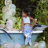 A young girl in a Meri Meri Mermaid Wrap Costume sits in a blue bathtub surrounded by lighted balloons and plush sea creatures, set against a backdrop of lush green foliage.